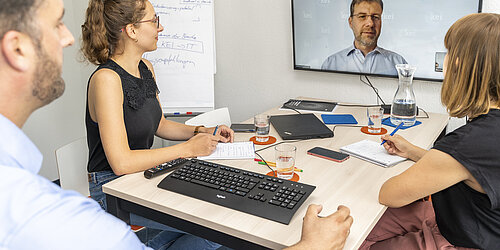 Drei Personen sitzen versammelt vor einem Videobildschirm im Rahmen einer Videokonferenz