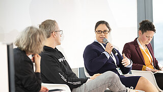 Eine Frau spricht in der Diskussionsrunde in ein Mikrofon während die anderen sie anschauen