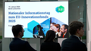 Titelfolie der Veranstaltung auf einem Screen, davor sitzen drei Personen