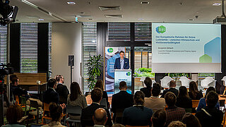Blick in den gefüllten Konferenzraum zum KEI Podium, auf der Bühne spricht Benjamin Görlach .