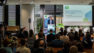 Blick in den gefüllten Konferenzraum zum KEI Podium, auf der Bühne spricht Benjamin Görlach .