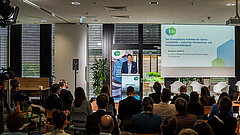 Blick in den gefüllten Konferenzraum zum KEI Podium, auf der Bühne spricht Benjamin Görlach .