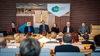 Sechs Personen sitzen vor einem gefüllten Hörsaal am Podium.