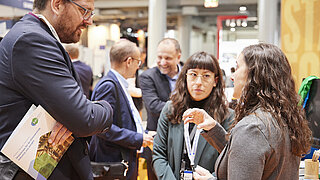 Zwei Mitarbeiterinnen des Kompetenzzentrums Klimaschutz in energieintensiven Industrien beraten einen Besucher am Messestand zur Hannover Messe.