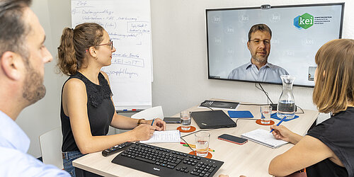 Drei Personen sitzen versammelt vor einem Videobildschirm im Rahmen einer Videokonferenz
