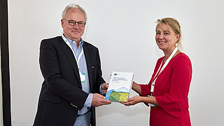 Eine Person überreicht einer anderen Person ein Buch mit freudigem Blick in die Kamera