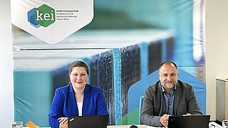 Die KEI Referent*innen Viktoria Metzler-Wowtscherk und Dr. Remo Tiedemann sitzen mit ihren Laptops an einem Tisch und begrüßen von dort die Teilnehmenden der Veranstaltung.