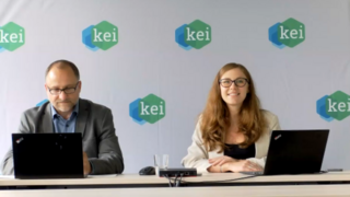 Zwei Personen sitzen vor jeweils zwei aufgeklappten Laptops vor einer Rückwand  