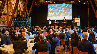 Blick in den Veranstaltungsraum zum German-Norwegian Energy Dialogue. Am Ende des Raumes ist eine Bühne aufgebaut auf der die Diskussionsteilnehmenden an Stehtischen stehen, vor der Bühne sitzen die Teilnehmenden an runden Tischen.
