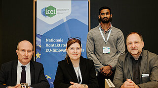 Drei Personen sitzen an einem Tisch. Ein vierte steht dahinter neben einem Roll-up mit dem Logo des KEI.