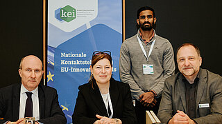 Drei Personen sitzen an einem Tisch. Ein vierte steht dahinter neben einem Roll-up mit dem Logo des KEI.