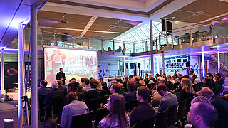 Blick auf die Bühne der Jahrestagung, im Vordergrund der gefüllte Veranstaltungsraum mit sitzenden Teilnehmenden.