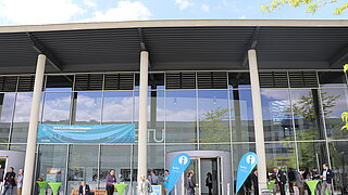 Circa 10 Personen stehen verteilt an bunten Stehtischen vor einem Universitätsgebäude