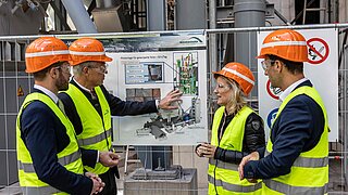 Bayrische Landtagsabgeordneter Daniel Artmann, Rosenheims Landrat Otto Lederer mit Daniela Ludwig, Parlamentarische Staatssekretärin beim Bundesminister des Innern, und Dr. Markus Stecher vom Rohrdorfer Net Zero Emission-Team schauen sich ein Schaubild der Anlage an. Alle Personen tragen Warnwesten und Schutzhelme.