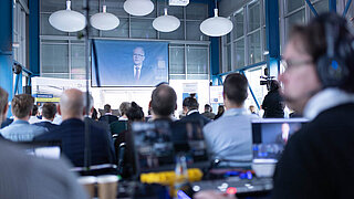 Blick von hinten in den gefüllten Veranstaltungsraum der Lausitzer Fachkonferenz 2023. Udo Philipp, Beamteter Staatssekretär beim Bundesminister für Wirtschaft und Klimaschutz (BMWK) richtet eine Video-Grußbotschaft an die Teilnehmenden.