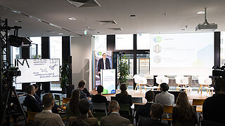 KEI-Leiter Jacob Flechtner steht auf der Bühne am Redner Pult, der Blick erfolgt von hinten aus dem Raum, man sieht das Publikum vor der Bühne sitzen. 