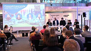 Die fünf Diskussionsteilnehmenden stehen an einem Stehtisch und sind im Gespräch. Im Hintergrund ist die Veranstaltungspräsentation zu sehen.