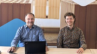 Die KEI Referenten Dr. Remo Tiedemann und Matthias Heck sitzen nebeneinander an einem Tisch und schauen in die Kamera. Vor ihnen steht ein Laptop.