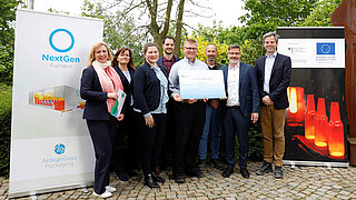 Acht Personen stehen aufgereiht vor Kamera und posieren mit dem Schild des Förderbescheids